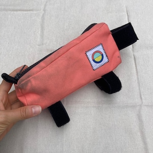 May include: A pink handlebar bag with black straps and a colourful circular logo. The bag has a zipper closure.
