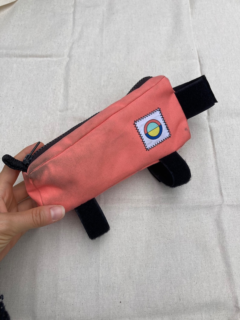 May include: A pink handlebar bag with black straps and a colourful circular logo. The bag has a zipper closure.