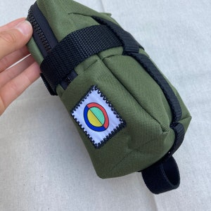 May include: A green canvas bike saddle bag with black straps and a zipper closure. The bag has a colorful circular patch with a white border.