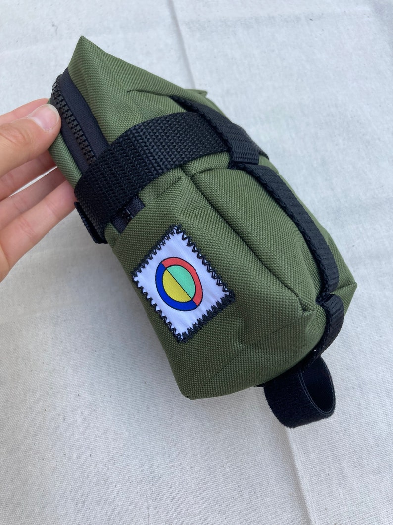 May include: A green canvas bike saddle bag with black straps and a zipper closure. The bag has a colorful circular patch with a white border.