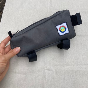 May include: Grey bike handlebar bag with a zip closure and a colourful circle logo patch. The bag has two black straps on the bottom and one black strap on the top.