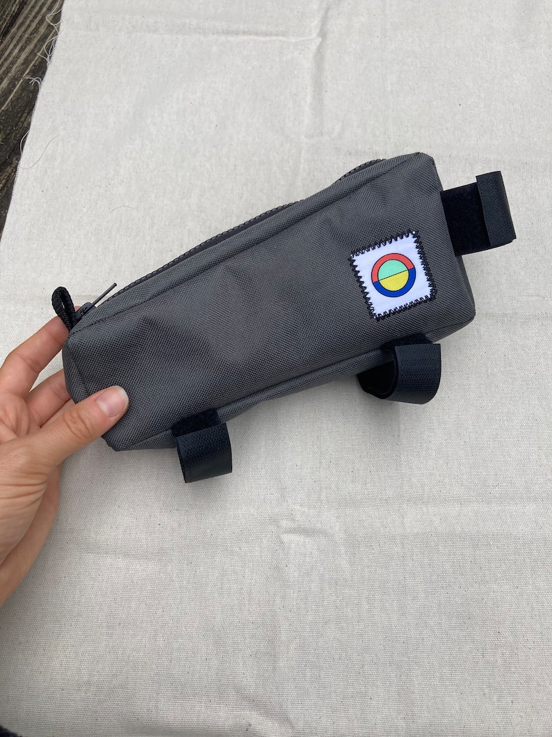 May include: Grey bike handlebar bag with a zip closure and a colourful circle logo patch. The bag has two black straps on the bottom and one black strap on the top.