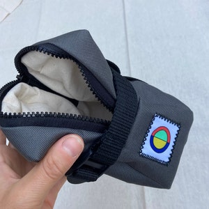 May include: A gray zippered bike saddle bag with a black strap and buckle. The bag has a small colorful logo with a circle and three colored segments.