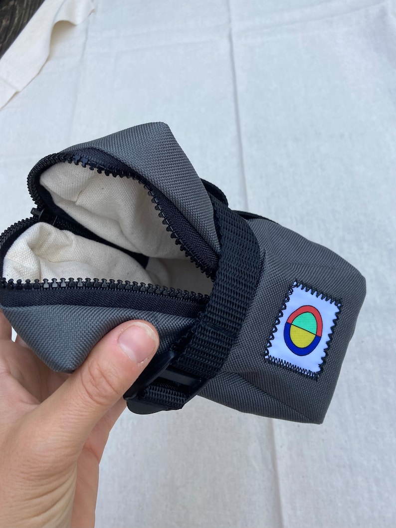 May include: A gray zippered bike saddle bag with a black strap and buckle. The bag has a small colorful logo with a circle and three colored segments.