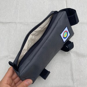 May include: A grey zipped bike handlebar bag with a black strap and a colourful circle logo. The bag is open and the inside is lined with a beige fabric.