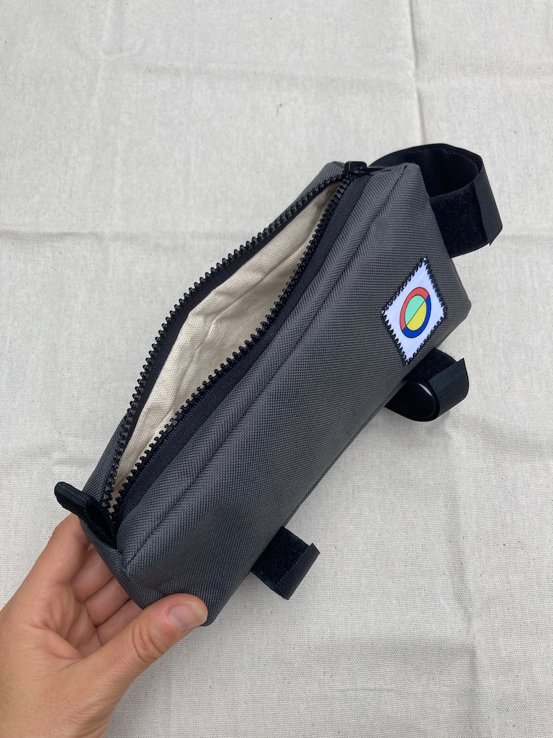 May include: A grey zipped bike handlebar bag with a black strap and a colourful circle logo. The bag is open and the inside is lined with a beige fabric.