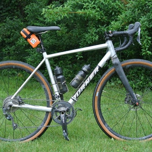 May include: A silver bicycle with brown tires and a black handlebar. The bicycle has two water bottle cages and a small orange bag attached to the seat post.