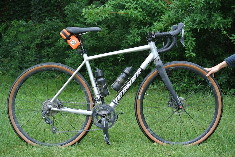 May include: A silver bicycle with brown tires and a black handlebar. The bicycle has two water bottle cages and a small orange bag attached to the seat post.