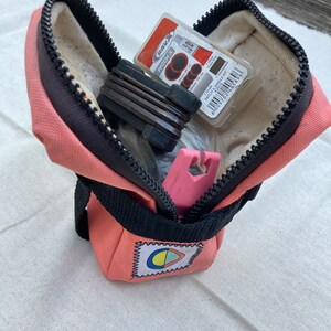 May include: A pink zippered pouch with a black strap and a colorful logo. The pouch is open and contains a set of bike tools, a patch kit, and a pink tire lever.