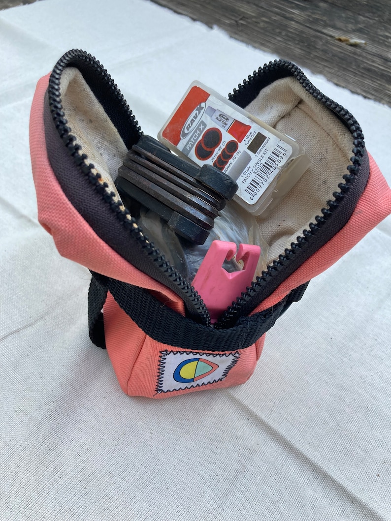 May include: A pink zippered pouch with a black strap and a colorful logo. The pouch is open and contains a set of bike tools, a patch kit, and a pink tire lever.