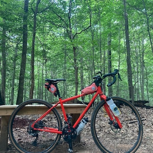 May include: A bright orange bicycle with black handlebars and wheels, parked on a wooden bench in a forest setting. The bike has two small orange bags attached to the frame and handlebars.