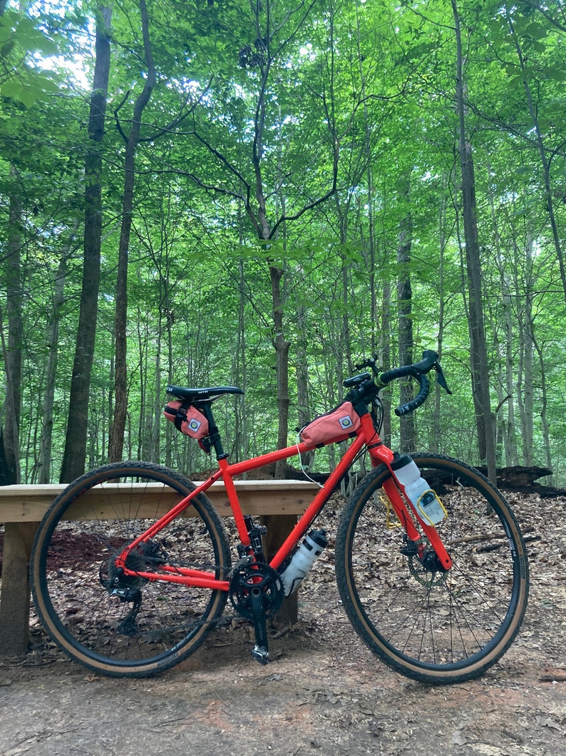 May include: A bright orange bicycle with black handlebars and wheels, parked on a wooden bench in a forest setting. The bike has two small orange bags attached to the frame and handlebars.