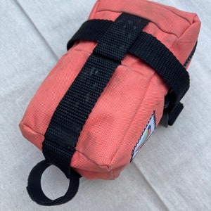 May include: A pink canvas bike bag with black straps and a zipper closure. The bag has a small colorful logo on the side.