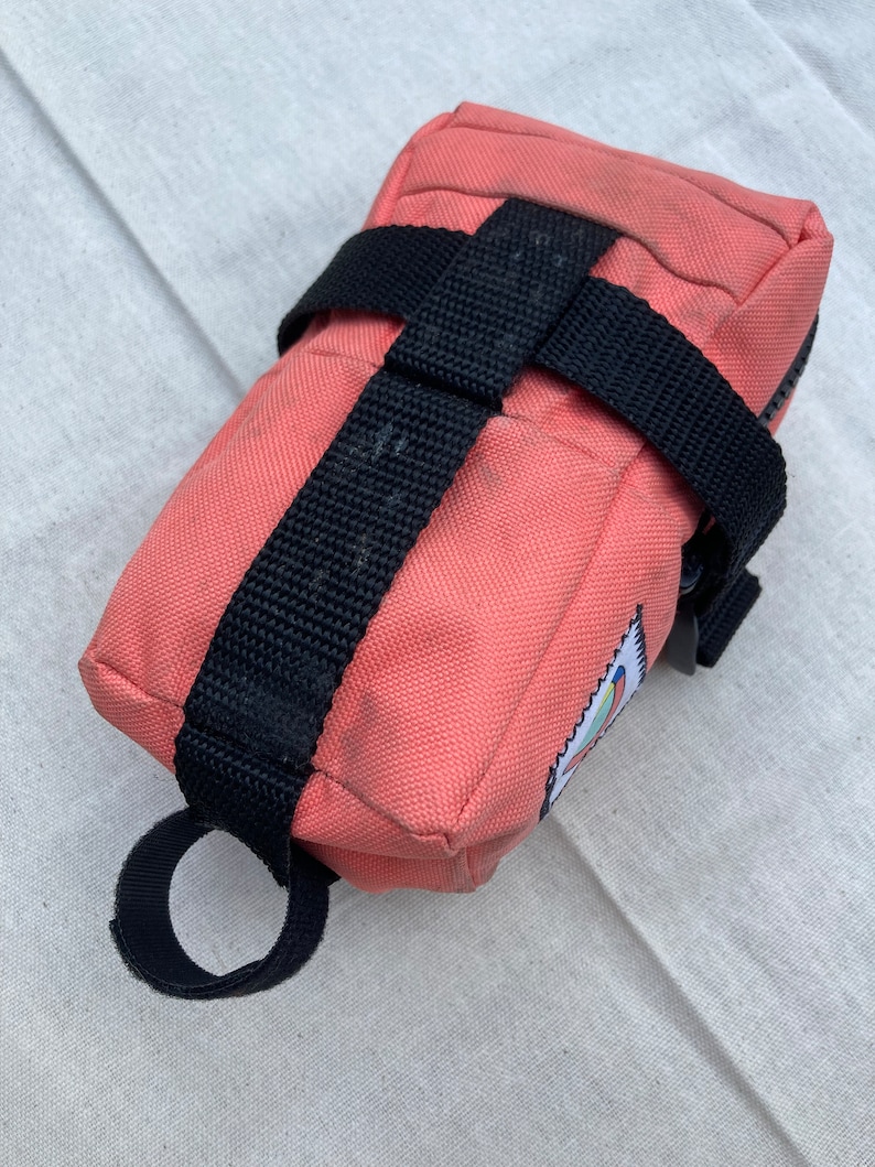 May include: A pink canvas bike bag with black straps and a zipper closure. The bag has a small colorful logo on the side.
