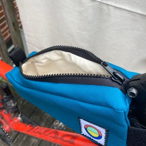 May include: A teal blue bike handlebar bag with a zipper closure and a small circular logo with red, yellow and green colours. The bag is attached to a red bicycle frame.