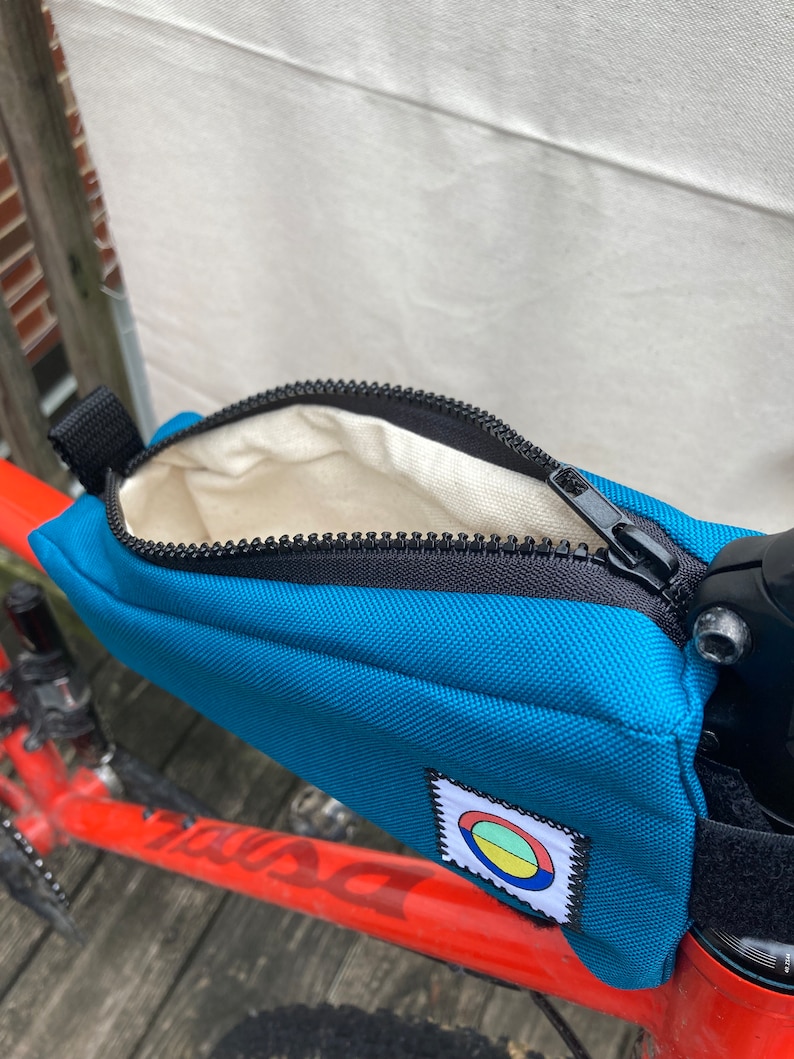 May include: A teal blue bike handlebar bag with a zipper closure and a small circular logo with red, yellow and green colours. The bag is attached to a red bicycle frame.