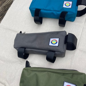 May include: Three different coloured bike handlebar bags. The bags are made of a canvas material and have a zipper closure. Each bag has a small patch with a colourful circle design on it. The bags are teal, grey, and green.