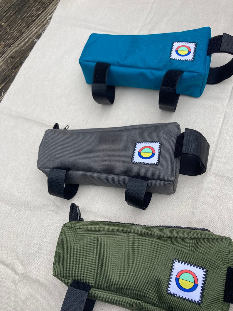 May include: Three different coloured bike handlebar bags. The bags are made of a canvas material and have a zipper closure. Each bag has a small patch with a colourful circle design on it. The bags are teal, grey, and green.