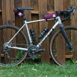May include: A silver and black bicycle with brown tyres and a burgundy handlebar bag. The bicycle is parked in front of a wooden fence.