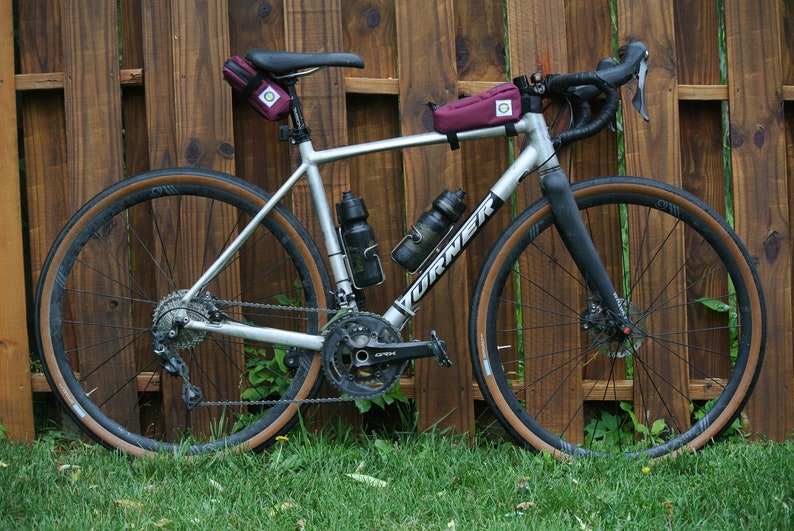 May include: A silver and black bicycle with brown tyres and a burgundy handlebar bag. The bicycle is parked in front of a wooden fence.