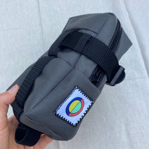 May include: Gray bike seat bag with a black strap and a colorful circle logo patch.