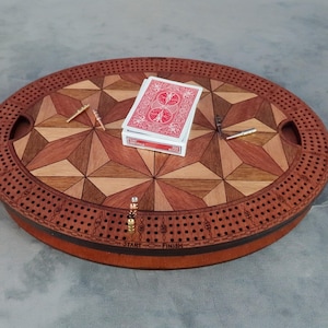 May include: Oval wooden cribbage board with a geometric inlaid design in shades of brown and tan. The board has a perimeter of holes for pegs, with a deck of cards and pegs resting on the surface. The game board has two handles.