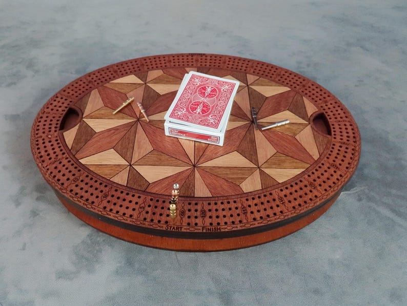 May include: Oval wooden cribbage board with a geometric inlaid design in shades of brown and tan. The board has a perimeter of holes for pegs, with a deck of cards and pegs resting on the surface. The game board has two handles.
