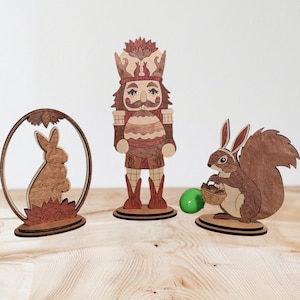 May include: Three wooden figurines: a nutcracker holding an egg, a rabbit, and a squirrel holding a basket. The nutcracker is wearing a brown and red outfit with a green hat. The rabbit is brown and the squirrel is brown with a bushy tail. The figurines are on a wooden surface.