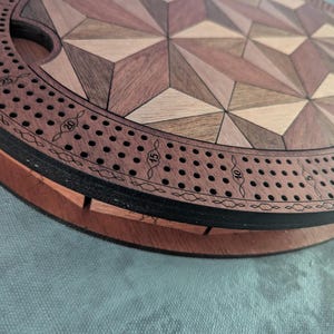 May include: A wooden cribbage board with a geometric design. The board features a circular track with numbered holes for pegs, and a central playing area with a starburst pattern in various wood tones. The board is approximately 12 inches in diameter.
