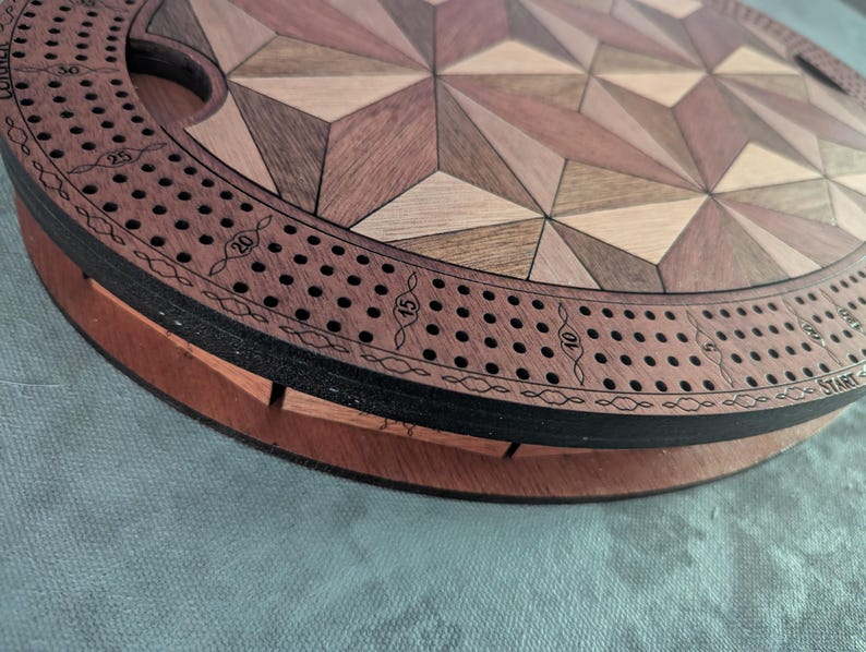 May include: A wooden cribbage board with a geometric design. The board features a circular track with numbered holes for pegs, and a central playing area with a starburst pattern in various wood tones. The board is approximately 12 inches in diameter.
