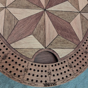 May include: Wooden cribbage board with a geometric starburst design in shades of brown and tan. The board has a circular border with numbered pegging holes and the word "SKUNK" engraved on the edge.