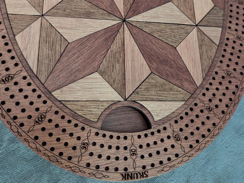 May include: Wooden cribbage board with a geometric starburst design in shades of brown and tan. The board has a circular border with numbered pegging holes and the word "SKUNK" engraved on the edge.
