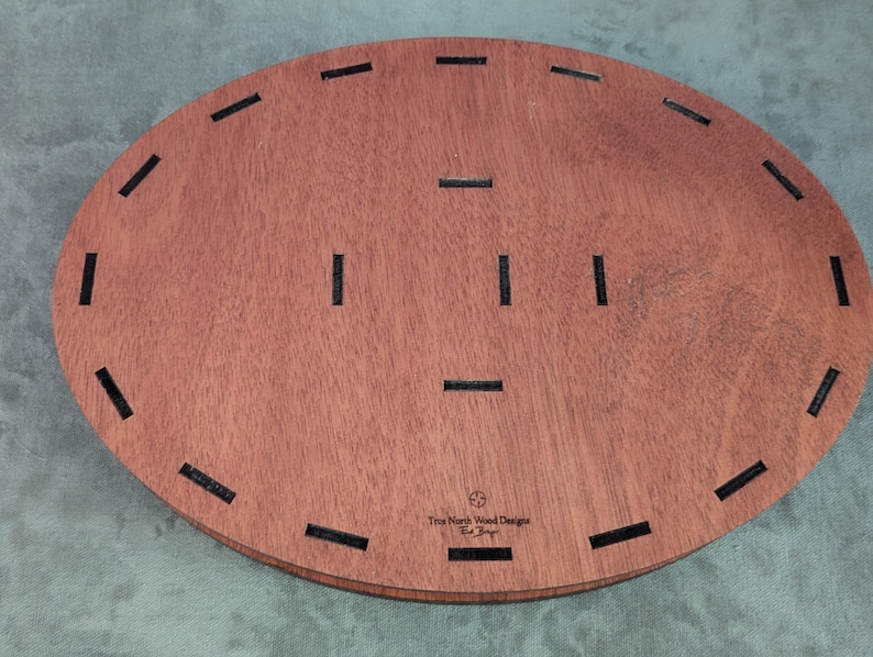 May include: Oval-shaped wooden tray with a reddish-brown finish. The tray has several evenly spaced, rectangular black inlays around the perimeter and in the center. The text "True North Wood Designs" is visible on the bottom right.