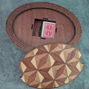 May include: A wooden cribbage board with a decorative geometric lid. The oval board is dark brown with drilled holes around the perimeter. A compartment holds a deck of cards and pegs. The lid features a starburst pattern in various wood tones.