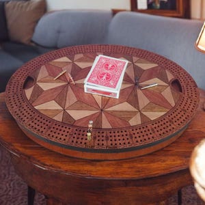 May include: A round wooden cribbage board with a geometric starburst design in shades of brown and tan. A deck of red playing cards and two pegs rest on the board. The board is on a wooden table.