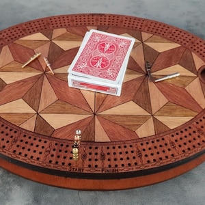 May include: A wooden cribbage board with a geometric design in shades of brown and tan. A deck of playing cards rests on the board, along with metal pegs. The board has a circular shape with a decorative border and handles.