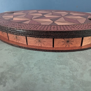 May include: A wooden cribbage board with a geometric design on the playing surface. The board is oval-shaped with a dark brown border and a light brown base. The base features floral engravings.