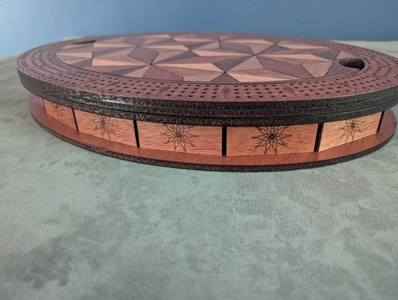 May include: A wooden cribbage board with a geometric design on the playing surface. The board is oval-shaped with a dark brown border and a light brown base. The base features floral engravings.