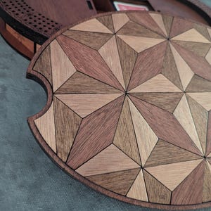 May include: A wooden game board lid with a geometric starburst pattern in shades of brown and tan. The lid is partially covering a round, wooden game board base. The design features a variety of wood tones and shapes.