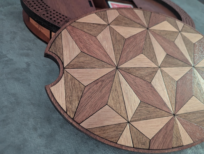 May include: A wooden game board lid with a geometric starburst pattern in shades of brown and tan. The lid is partially covering a round, wooden game board base. The design features a variety of wood tones and shapes.