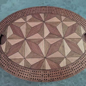 May include: Oval wooden cribbage board with a geometric starburst design in shades of brown and tan. The board has a track for pegs and cut-out handles. The words "CORNER", "SKUNK", "START", and "FINISH" are visible.