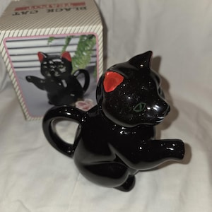 Puede incluir: Una tetera de cerámica negra con forma de gato, con detalles de orejas rojas y ojos verdes. La tetera es brillante y tiene un asa y un pico. En el fondo hay una caja con las palabras "BLACK CAT", que muestra una imagen de la tetera.
