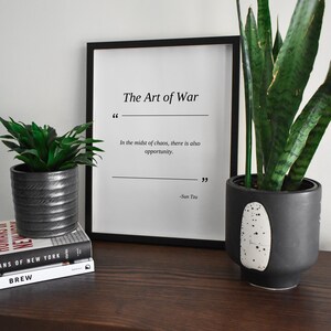 May include: A black framed print with the text "The Art of War" and a quote by Sun Tzu: "In the midst of chaos, there is also opportunity." The print is sitting on a wooden table with two potted plants.