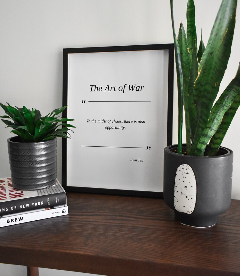 May include: A black framed print with the text "The Art of War" and a quote by Sun Tzu: "In the midst of chaos, there is also opportunity." The print is sitting on a wooden table with two potted plants.