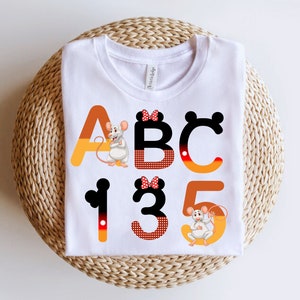 May include: White t-shirt with a black, red, and yellow alphabet design featuring a mouse character. The letters are A, B, C, 1, 3, and 5.