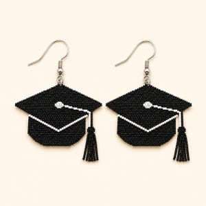May include: A pair of beaded earrings shaped like graduation caps. Each earring features a black square mortarboard with a white tassel and a white button detail at the center. The earrings are attached to silver-toned metal fishhook ear wires.
