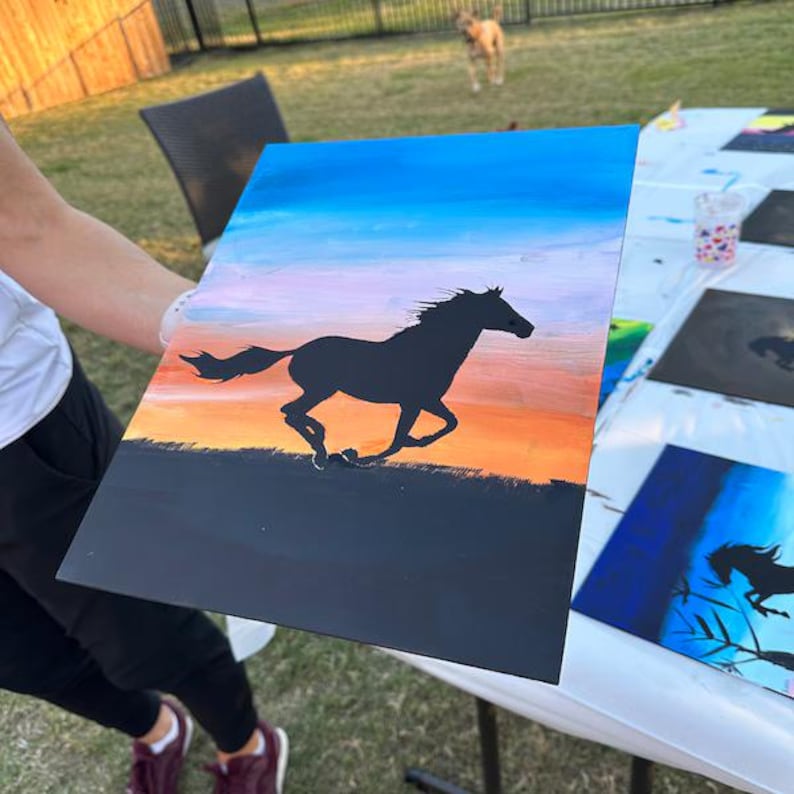 May include: A rectangular painting on a flat surface featuring a black silhouette of a horse galloping across a landscape. The background displays a gradient sky transitioning from blue at the top to light pink and vibrant orange near the horizon line.