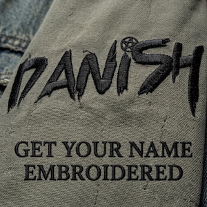May include: An olive green fabric patch featuring the black embroidered text DANISH with a small pentagram symbol above the letter I. Below this, the text GET YOUR NAME EMBROIDERED is stitched in a clean, black, sans-serif font on the canvas.