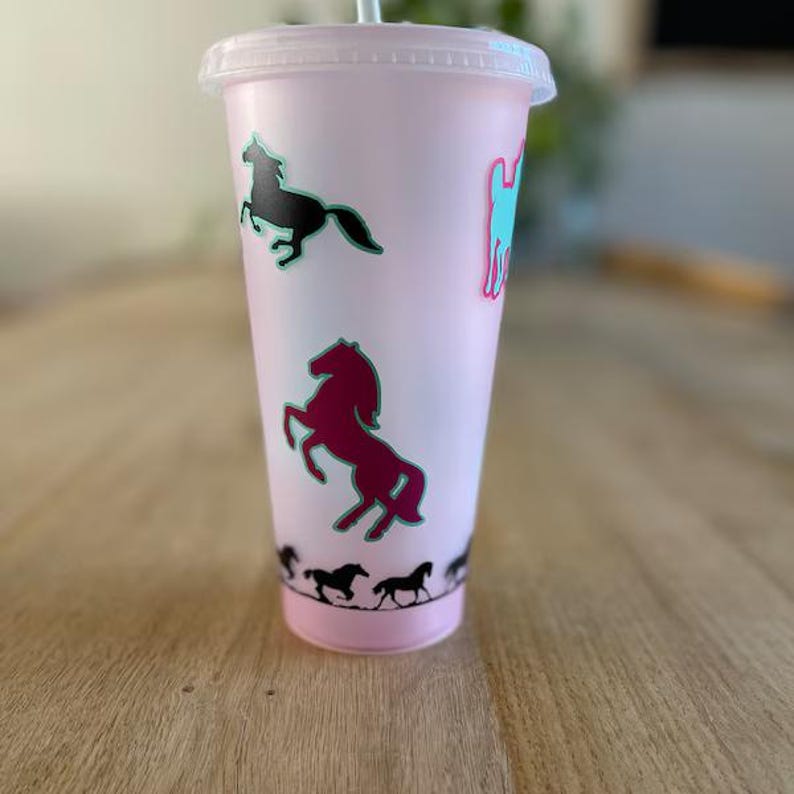 May include: A light pink plastic tumbler with a clear lid and a straw, featuring various horse silhouettes in black, dark red, and teal. The cup displays a large rearing horse in dark red, a running horse in black, and a border of small horses.