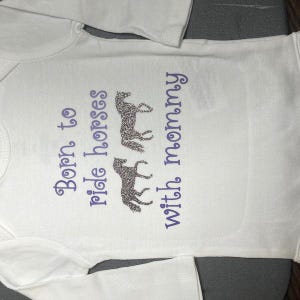 May include: A white long-sleeved baby bodysuit featuring the purple text Born to ride horses with mommy and two glittery silver horse silhouettes positioned between the words ride and horses and the word with.